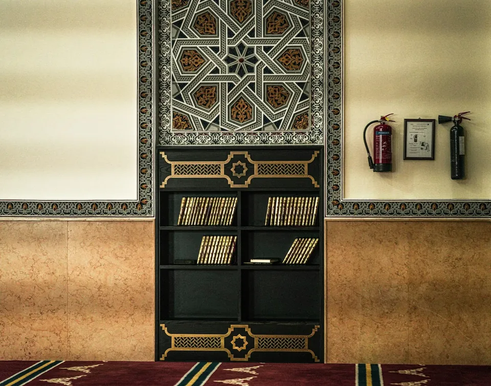 جدار يعلوه لوح مزخرف بنقش نجمي هندسي فوق خزانة كتب داكنة، بجوار طفايات حريق على سجادة حمراء.