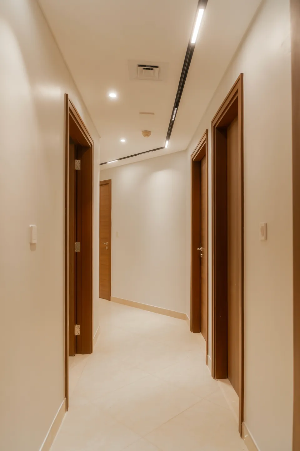 Imperial Avenue Apartment, Downtown Dubai: cream hallway with wooden door frames, recessed lights and a linear ceiling light.