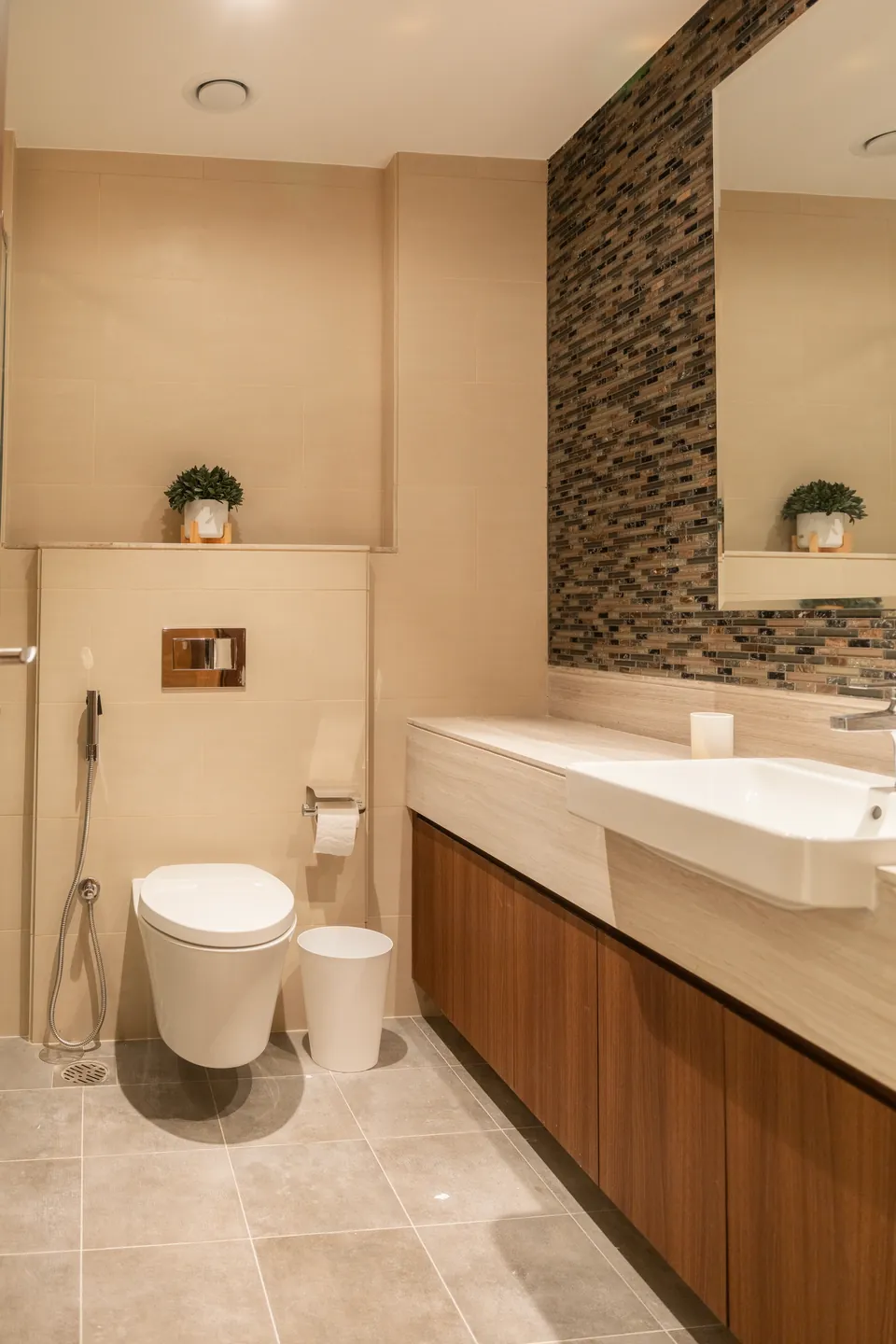 Imperial Avenue Apartment, Downtown Dubai: bathroom with a wall-hung toilet, wood vanity, white basin and mosaic tiles.