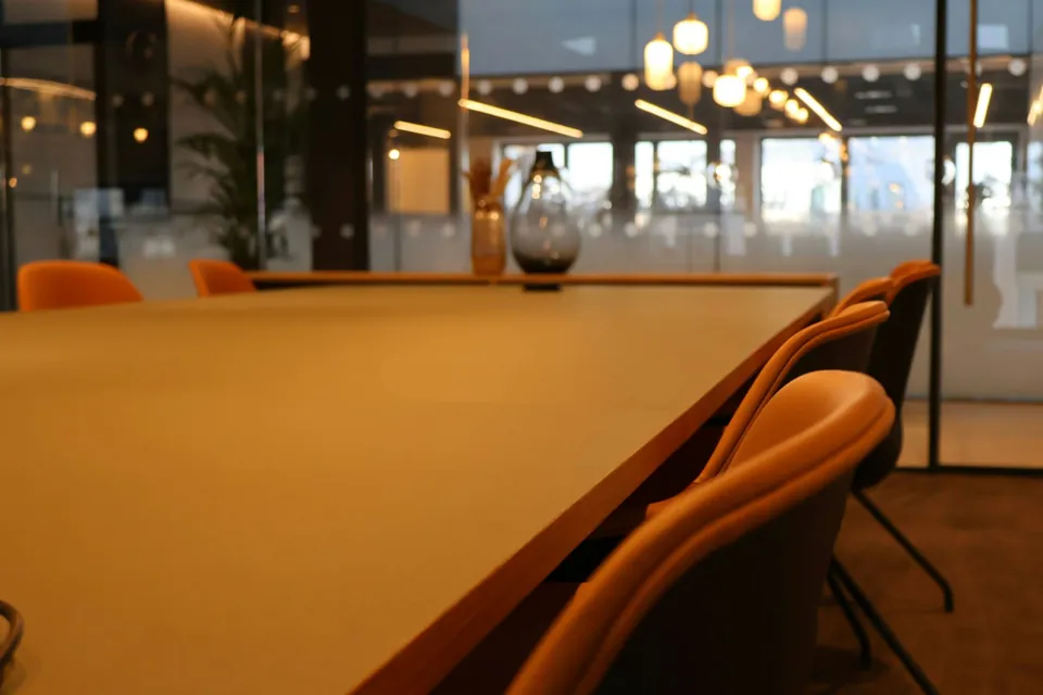 طاولة اجتماعات بكراسٍ برتقالية في مكتب بجدران زجاجية تضيئه مصابيح معلّقة.